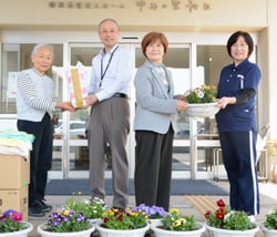 土屋常務（左から二番目）と山﨑施設長（右）に鉢花と清拭布を渡す、森田支部長（右から二番目）ら