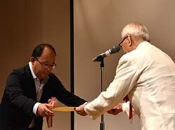 功績を称え記念品を渡す河崎委員長（右）と藤本社長（中央）
