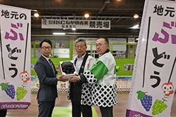 堀社長（左）に「デラウェア」を手渡す田中組合長（中）と上條常務理事