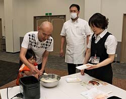 岡崎係長(中央)にアメリカンドックの作り方を習う親子（左）