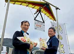 スタート地点をバックにゴール地点で幻の米をPRする湯本正行部会長と柳武雄さん