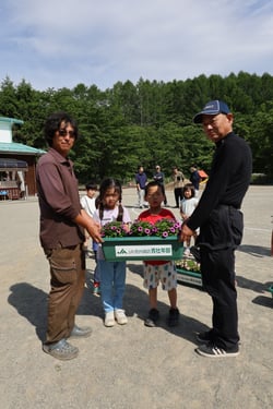 原村保育所の園児にプランターを渡す清水ブロック長（左）と野明輝彦副ブロック長（右）（長野県原村で）