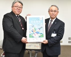 橋渡教育長に冊子のパネル版を手渡す相馬常務㊧（長野県安曇野市で）