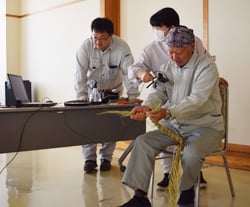 わらを編む伊東さんの手元を撮影する職員